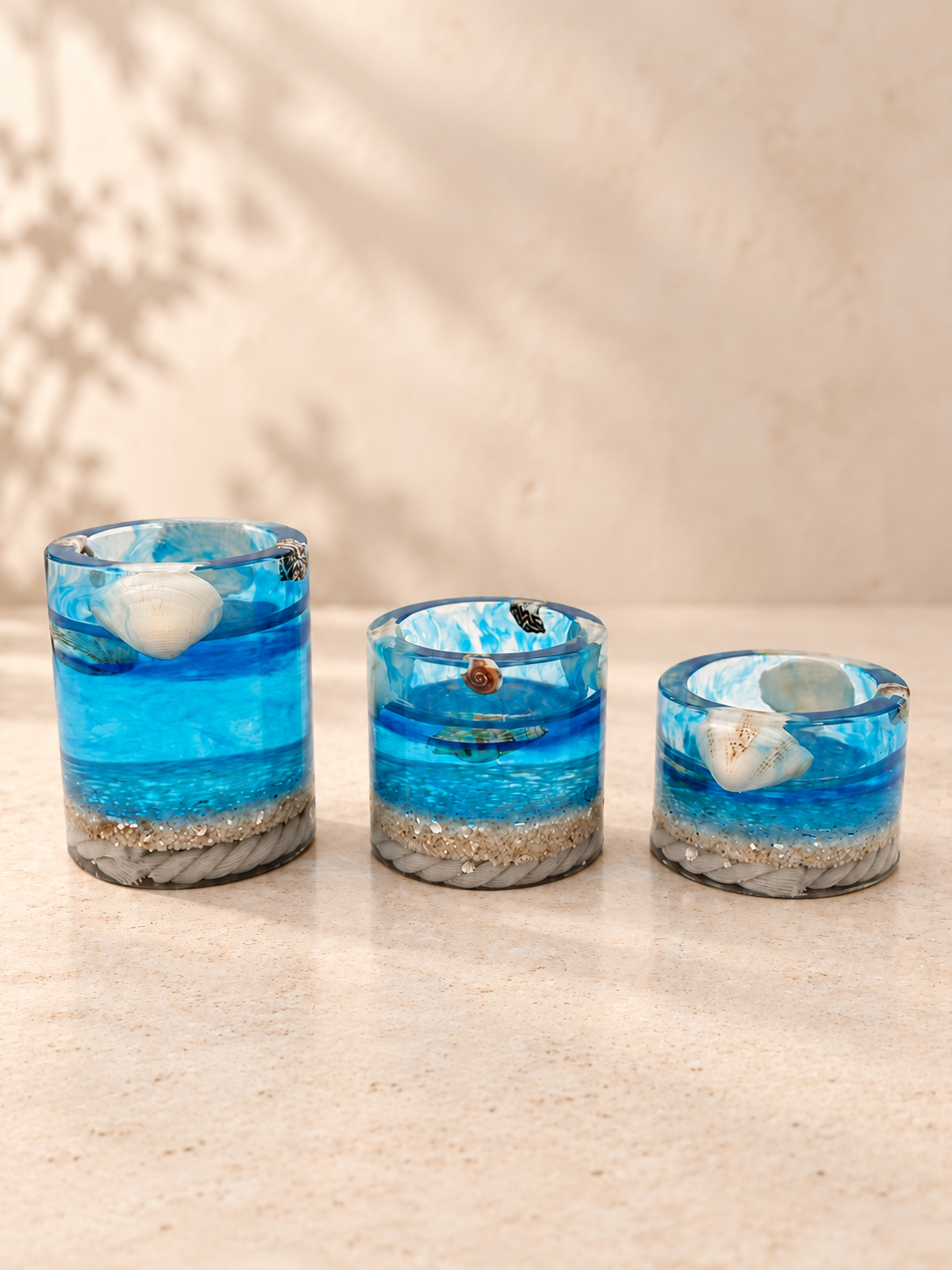 Set of three graduated ocean-blue resin candle holders with shell details on an ivory tabletop, handmade in Lancashire UK by Held Forever Studio