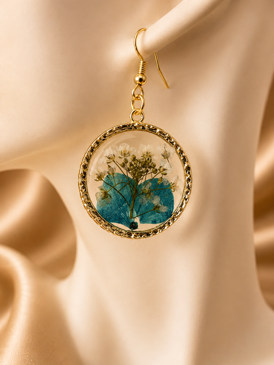 Close up of pressed flower resin earring showing blue hydrangea and gypsophila detail in UV resin with gold circular bezel frame, handmade by Held Forever Studio