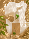 Botanical resin drop earrings with real pressed green leaves and white dried flowers in gold-tone rectangular frames, displayed on kraft earring card — handmade by Held Forever Studio in Lancashire, UK