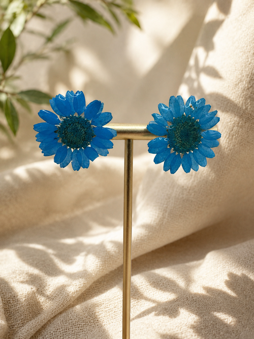 Blue daisy resin stud earrings on gold display stand with greenery and dried flowers, handmade in Lancashire UK