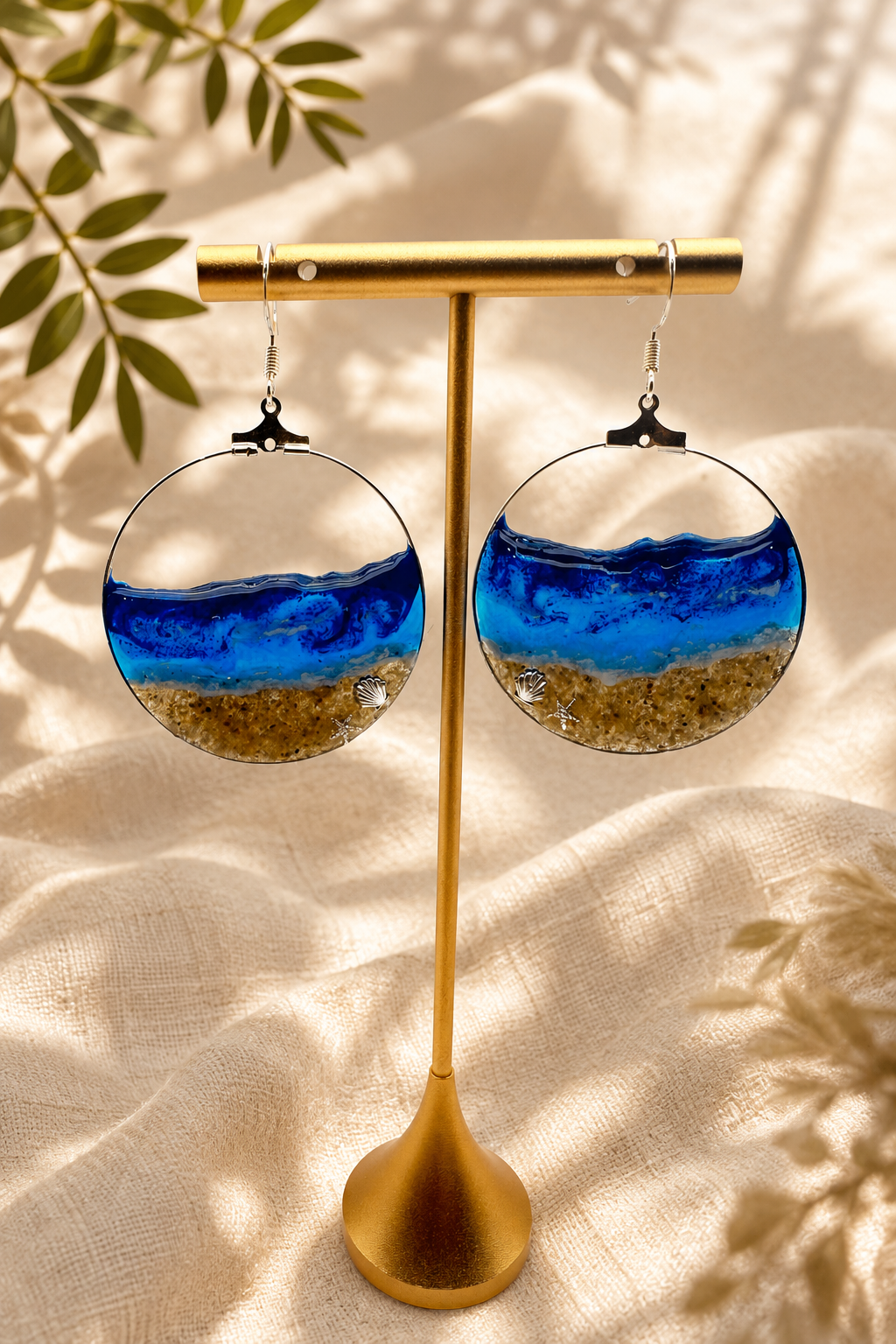 Pair of ocean wave resin hoop earrings on a gold display stand, layered blue and white resin over sand with silver shell and starfish charms, handmade by Held Forever Studio